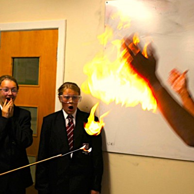Two schoolgirls in uniform react with amazement and surprise as a teacher demonstrates a flaming hands science experiment in a classroom, with visible flames and safety goggles worn for protection.
